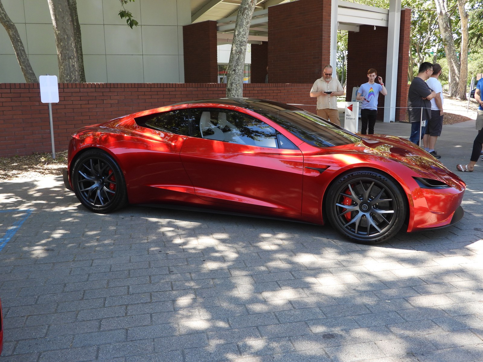 The red second-generation Tesla Roadster prototype parked outdoors on a paved plaza in daylight, trees casting shadows across the car and visitors standing behind a barrier